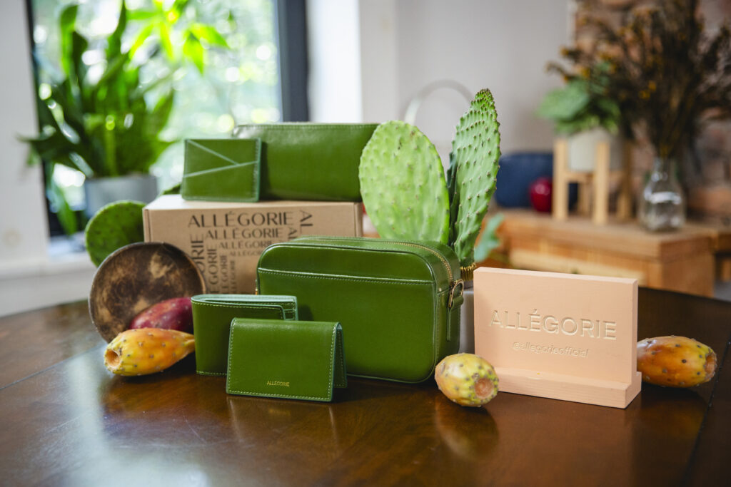 Allegorie's sustainable accessories - including wallets, purses, and bags - sit on a brown table surrounded by cactus.