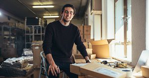 A business owner who knows how to calculate break even point sits at a desk surrounded by shipping boxes.