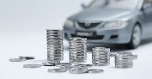 Several stacks of coins in smaller and smaller groups demonstrating the concept of depreciation. In the background of the photo a silver sedan car is parked.